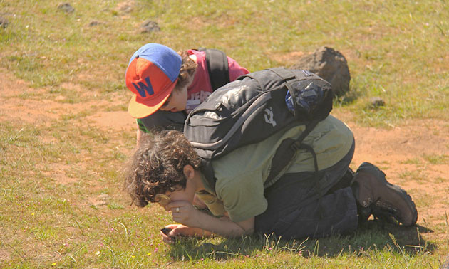 Kids crouched on ground examining something with magnifying glass. 
