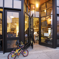 A welcoming entrance comprised of glass windows lookin on a warm-lit scene and diners at tables. Two small bikes stand outside. 
