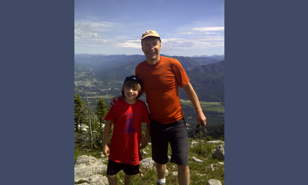 A man and young boy stand together on top of a mountain in summer