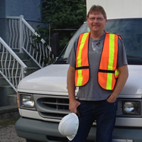 Max stands in front of a white cube van wearing a safety vest and holding a hard hat.