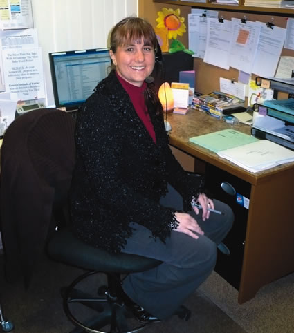 A smiling woman sits at an office desk with a phone headset on.