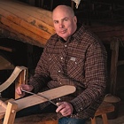 Photo of Mike Elliott working on a wooden canoe paddle with wood canoes behind