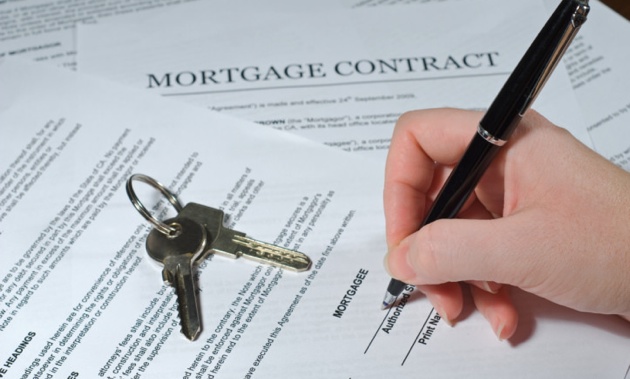A photo of a hand signing a mortgage document.  House keys are sitting on the document. 