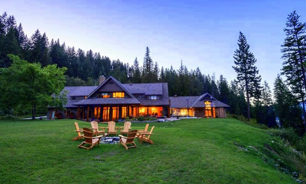 Picture of log-cabin style lodge house, with Adirondack chairs sitting on the lawn, and a treed mountainside in the background. 