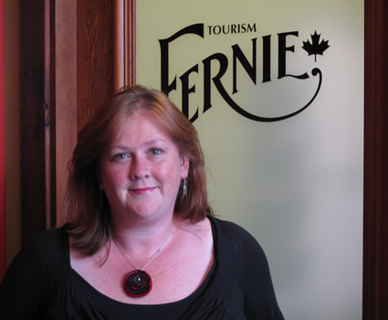 Photo of a brown haired lady in a black shirt standing in front of a door that says Tourism Fernie on it. 