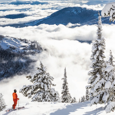 Picture of skier standing on mountainside with view of distant mountains and clouds. 