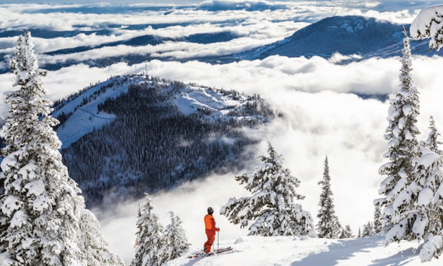 Picture of skier standing on mountainside with view of distant mountains and clouds. 