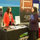 A woman in a red blazer behind a Lethbridge College table communicates with a student in a purple coat.