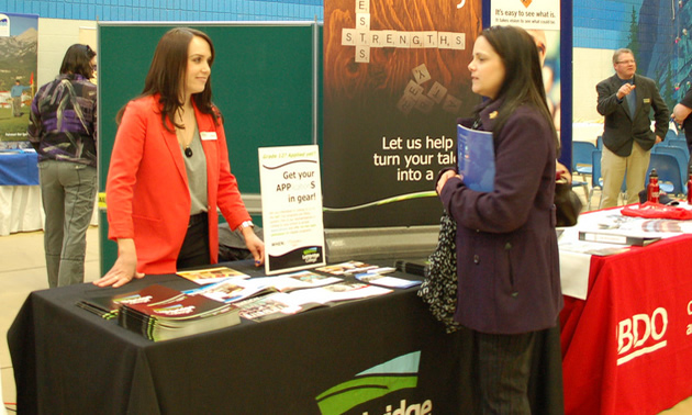 A woman in a red blazer behind a Lethbridge College table communicates with a student in a purple coat.