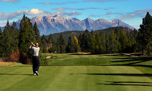 Golfer taking a swing along a long fairway, with large mountains in background. 