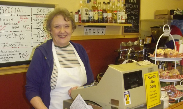 a middle-aged woman smiling behind a counter with an apron on and whiteboard with daily specials behind