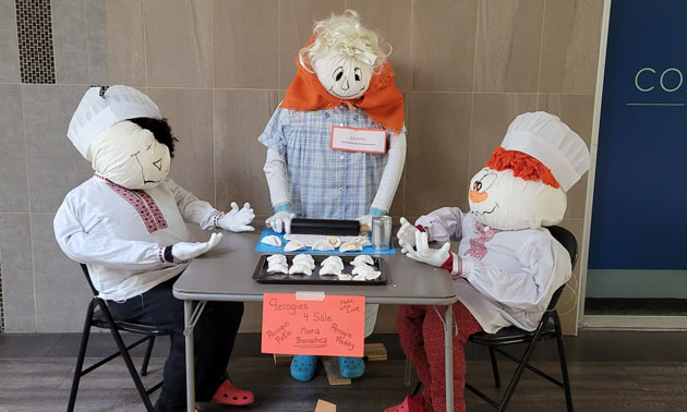 Creative scarecrow display showing three scarecrows seated around small card table, making perogies. 