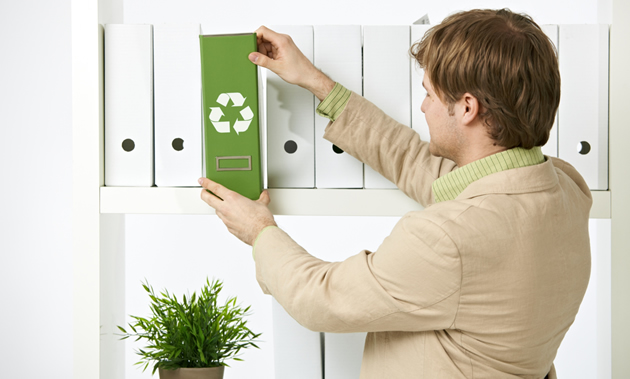 A man in a suit jacket pulls a green binder with a recycle symbol off of a shelf of white binders.