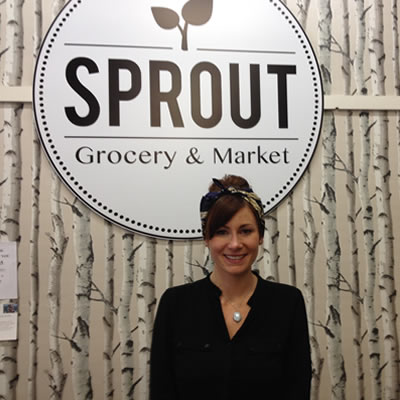 Chantel Delaney is standing in front of the Sprout Grocery sign with birch tree wallpaper on the wall.