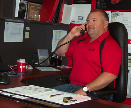Photo of a business man in a red shirt talking on the phone at his desk. 