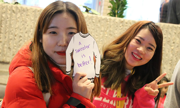 Two female students are wearing warm layers of clothes and are holding a sign that says 