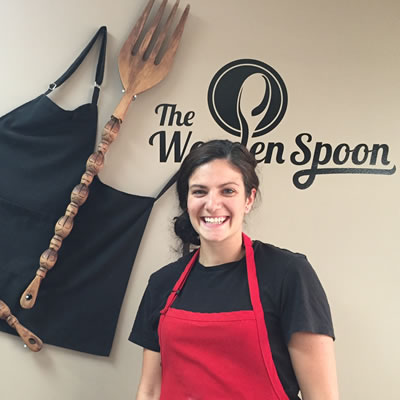 Owner and chef Kayla Sebastian of The Wooden Spoon Bistro & Bake Shop in Grand Forks is standing in front of the bistro's logo.