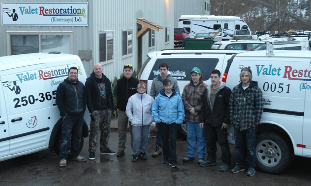 A group of nine people wearing jeans, jackets and work boots stand outdoors near vehicles carrying the logo Valet Restoration Services Ltd.