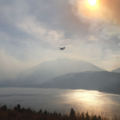 A water bomber is flying over Moyie Lake with the sun filtered by smoke.