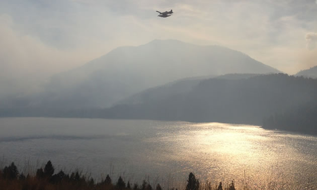 A water bomber is flying over Moyie Lake with the sun filtered by smoke.