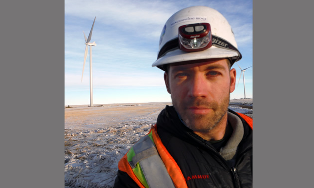 Randolph Seibold wears a hard hat and headlamp. He's standing next to a wind tower in a snowy landscape.