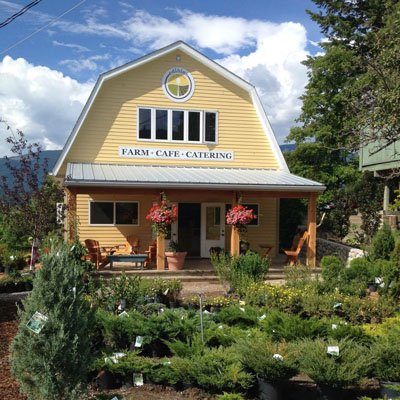 Barn-roofed building, butter-yellow in colour with white trim, deck out front, plants in foreground. 