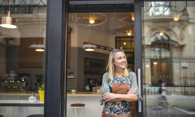 Young restaurant owner leaning in doorway with arms crossed, smiling. 